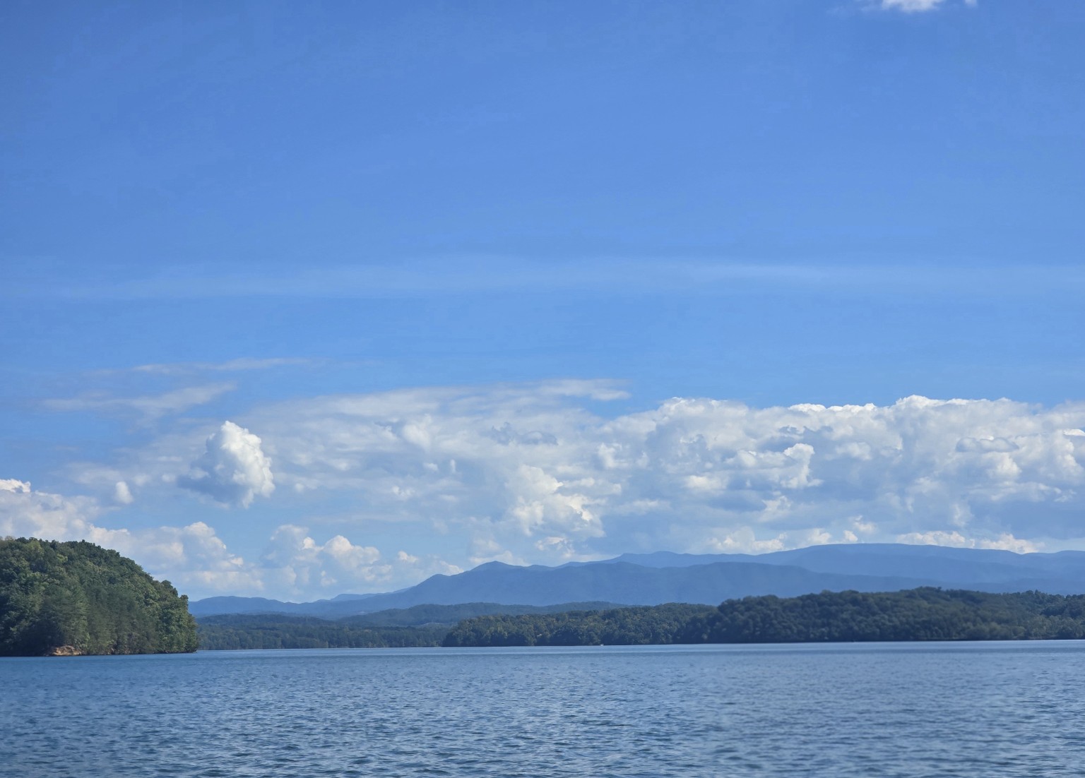 A wide view across blue water toward wooded hills and mountains beneath a broad sky
